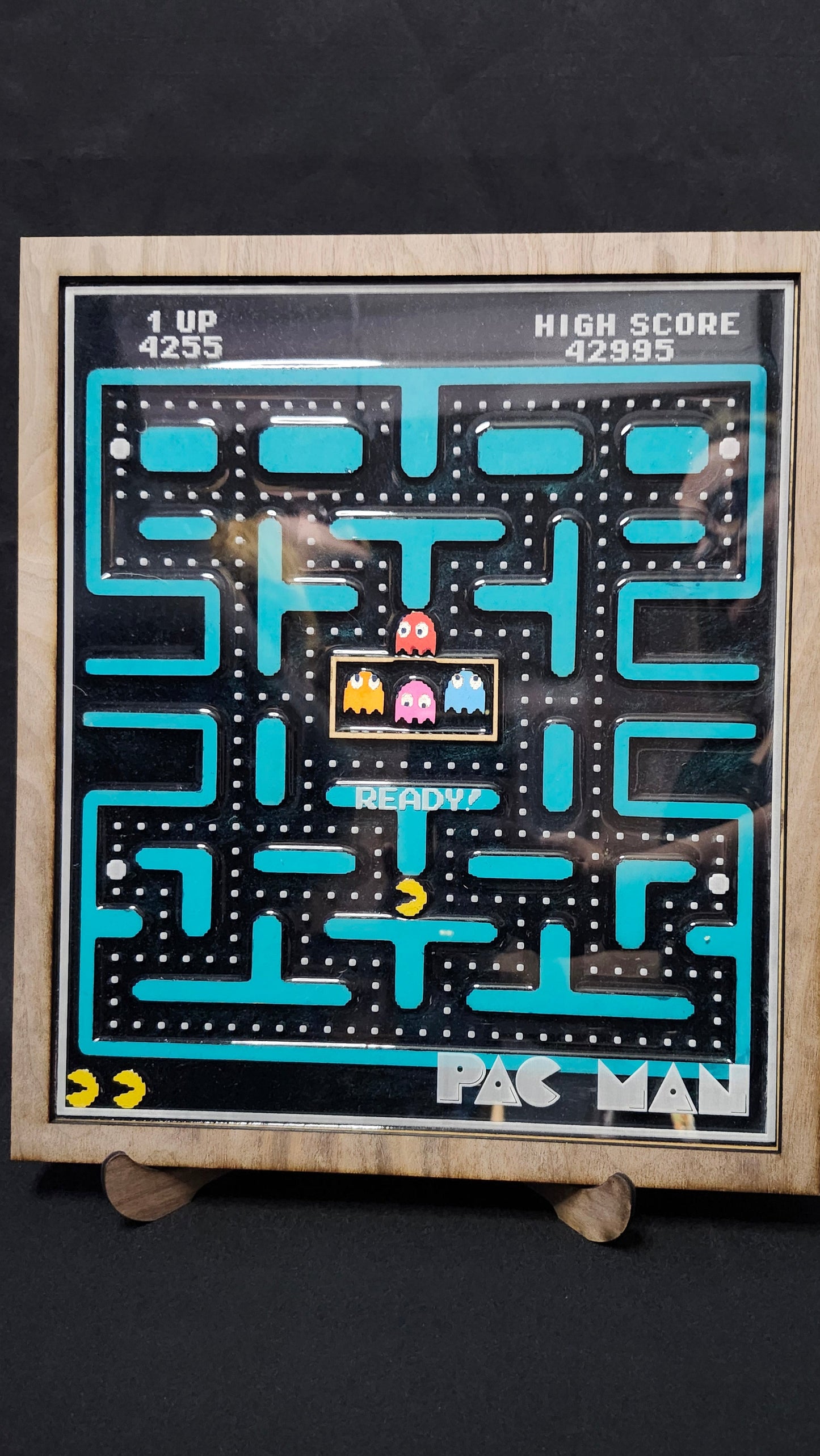 A framed retro Pac-Man arcade board game featuring the classic maze with blue paths, a yellow ghost, and red and pink ghosts, set against a dark background.