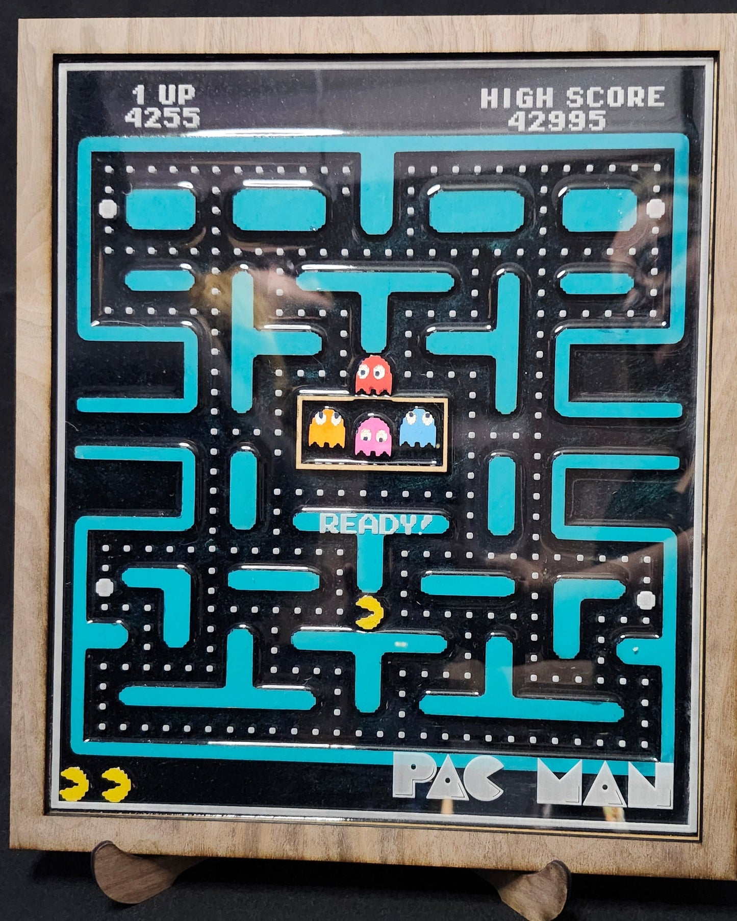 A framed retro Pac-Man arcade board game featuring the classic maze with blue paths, a yellow ghost, and red and pink ghosts, set against a dark background.