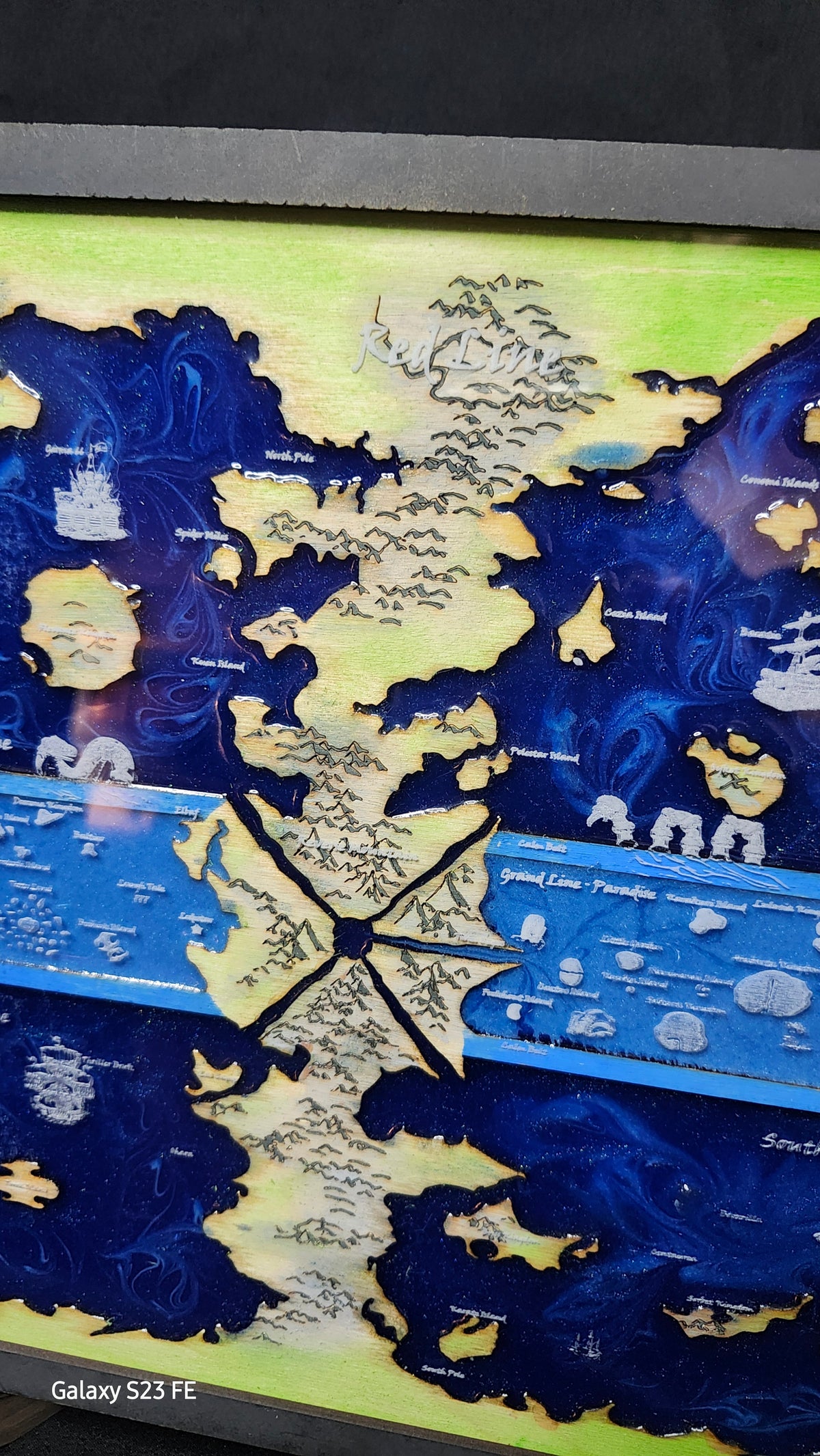 A close-up of a hand painted map art piece featuring fantasy landscapes in deep blue and light green, with white illustrated details and text labels, photographed indoors.
