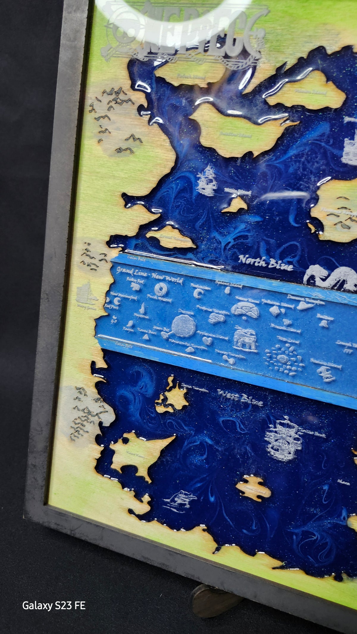 A close-up of a hand painted map art piece featuring fantasy landscapes in deep blue and light green, with white illustrated details and text labels, photographed indoors.
