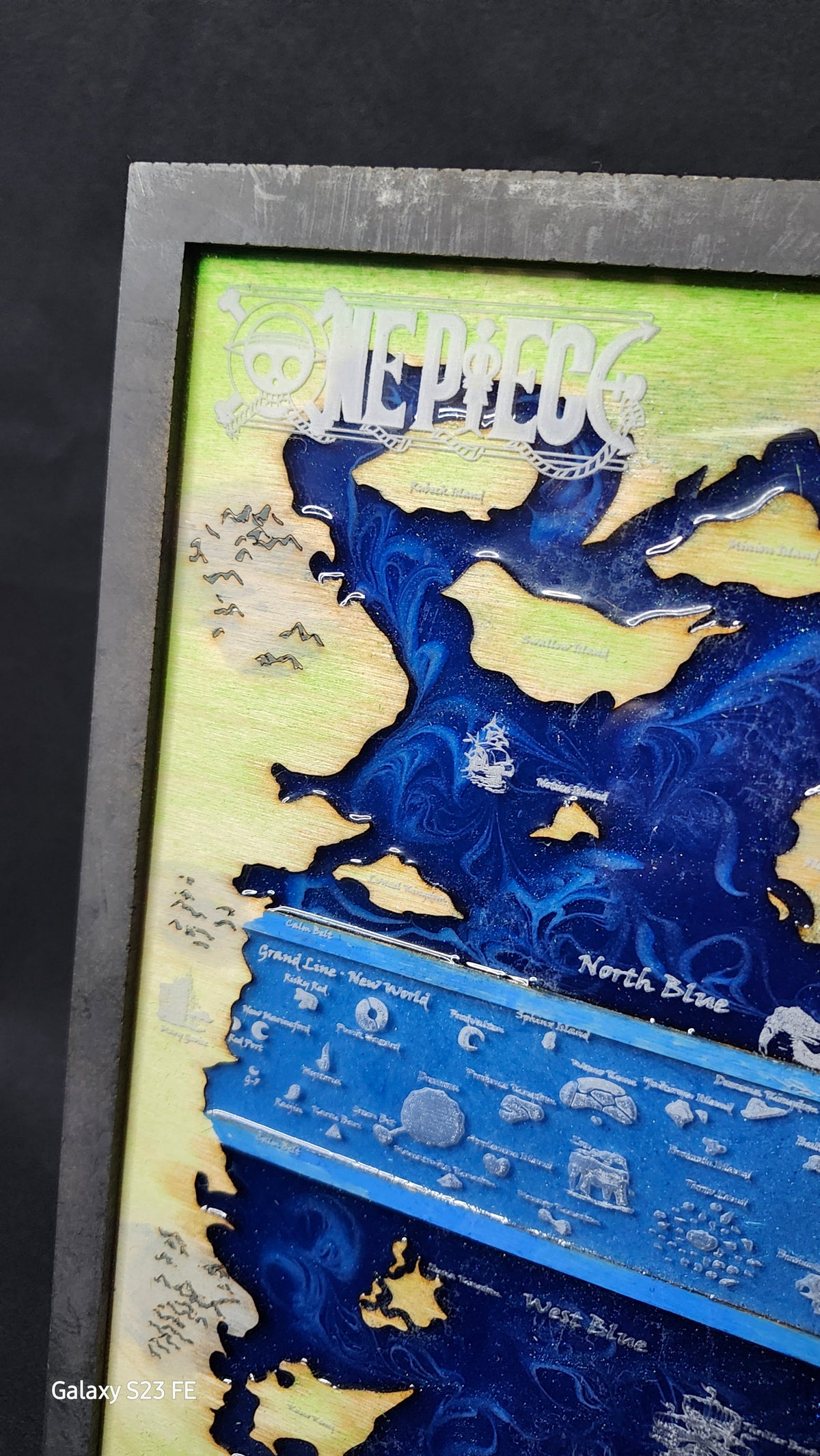 A close-up of a hand painted map art piece featuring fantasy landscapes in deep blue and light green, with white illustrated details and text labels, photographed indoors.