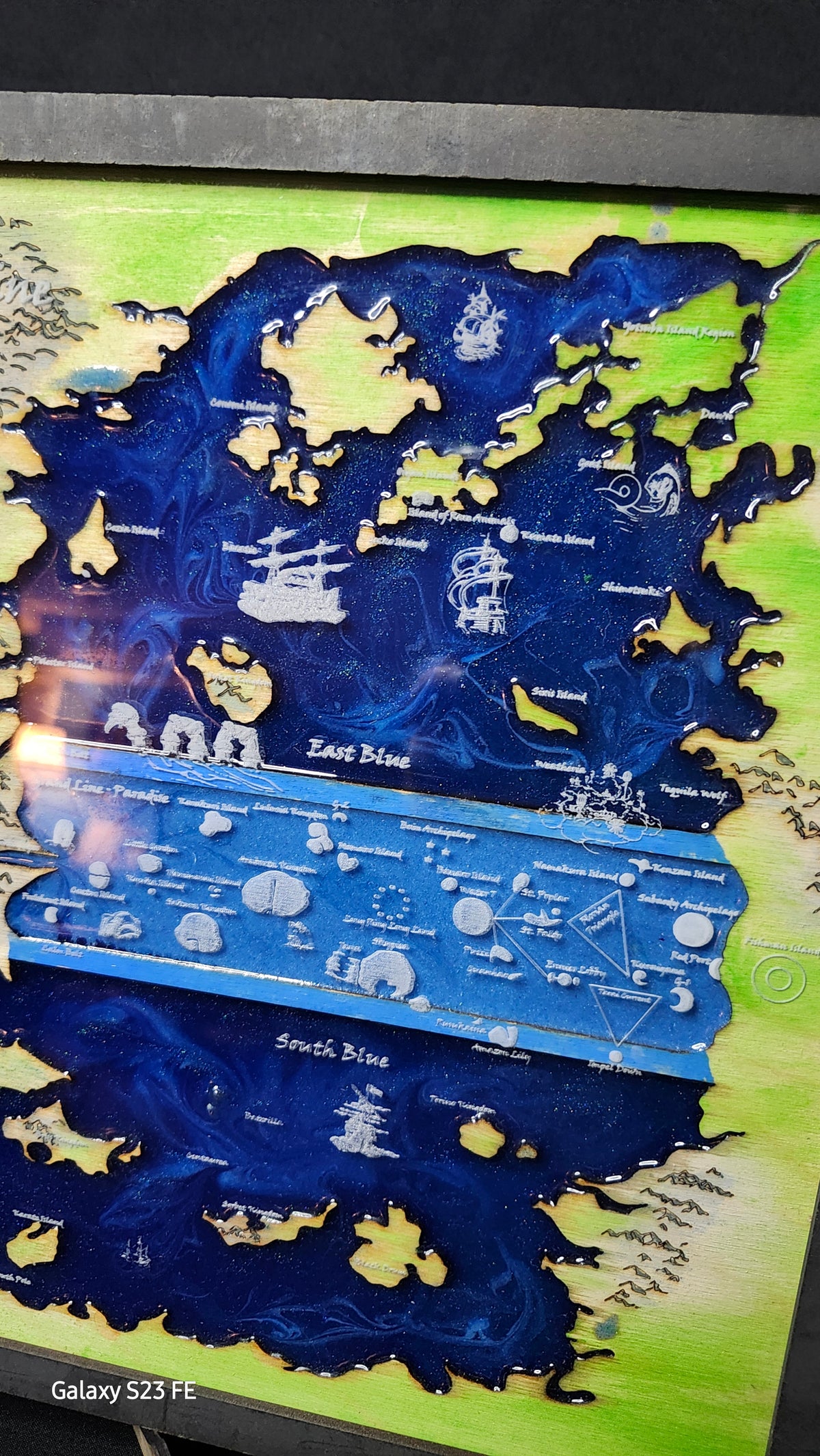 A close-up of a hand painted map art piece featuring fantasy landscapes in deep blue and light green, with white illustrated details and text labels, photographed indoors.